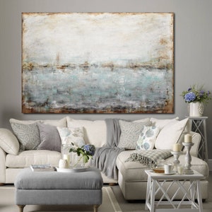 May include: A large abstract painting with a white background and a horizontal band of blue, green, and brown paint. The painting is hanging on a wall above a white sectional sofa with gray throw pillows and a gray ottoman.