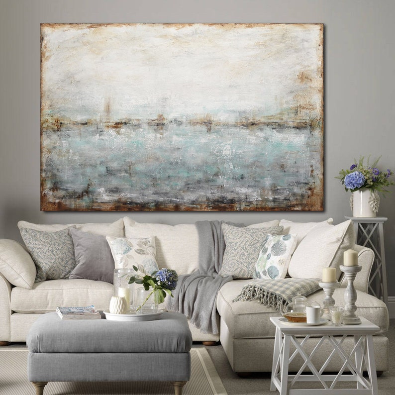 May include: A large abstract painting with a white background and a horizontal band of blue, green, and brown paint. The painting is hanging on a wall above a white sectional sofa with gray throw pillows and a gray ottoman.