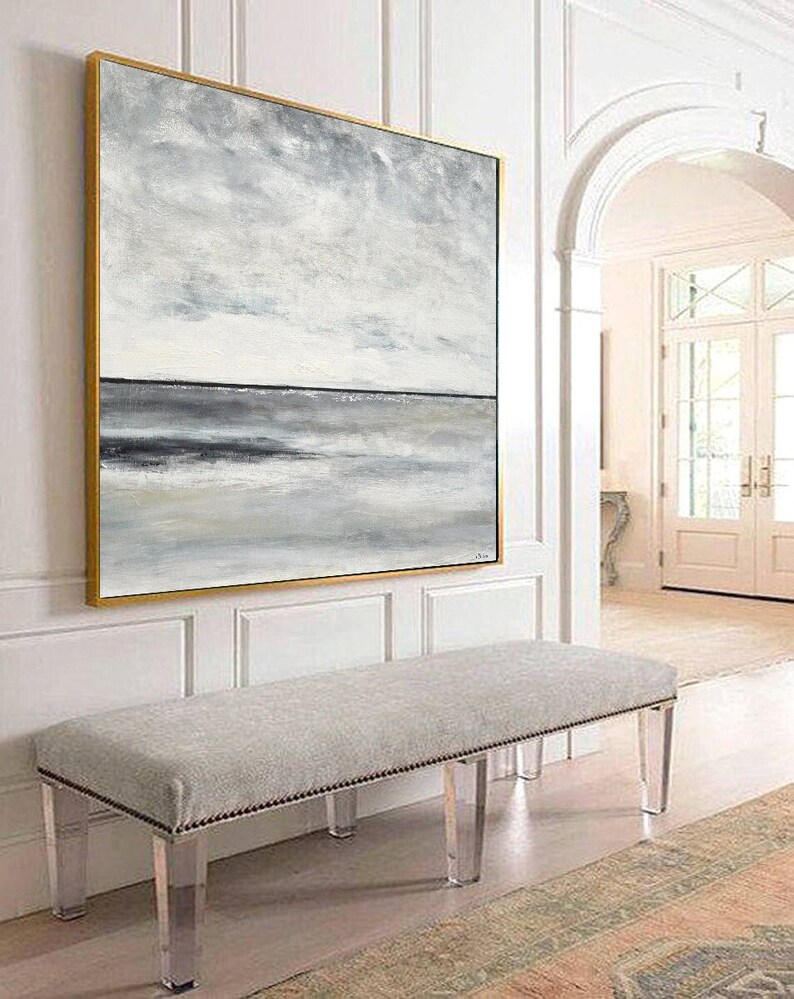 May include: A large abstract painting of a seascape in shades of gray and white, framed in gold, hanging on a white wall above a gray upholstered bench with clear acrylic legs.