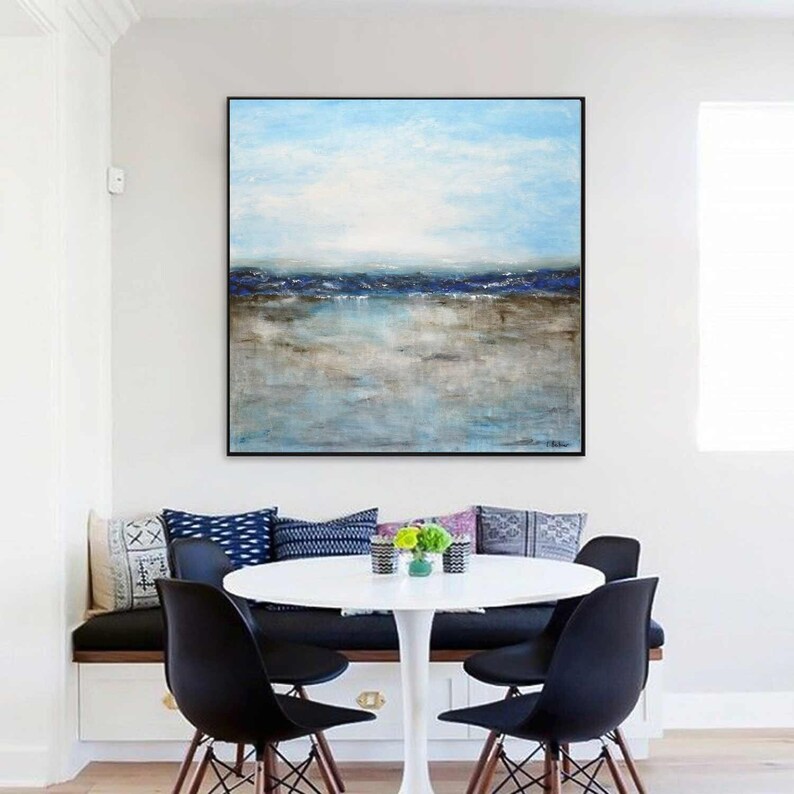 May include: Abstract painting of a seascape with blue and brown hues. The painting depicts a horizon with a blue sky and a body of water with a sandy shore. The painting is framed in black and hangs on a white wall above a dining table.