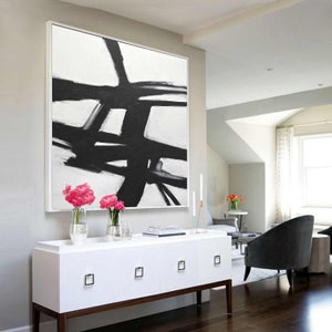 May include: A large black and white abstract painting with bold brushstrokes hangs above a white dresser with four drawers. The painting is framed in a thin white frame.