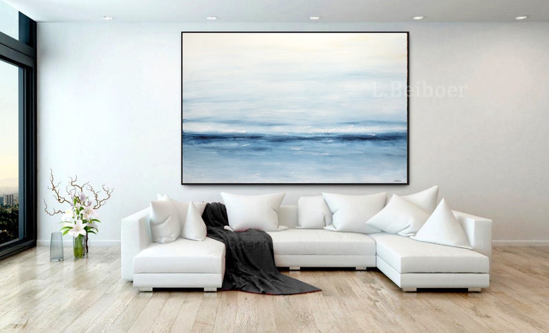 May include: A large abstract painting of a seascape in shades of blue and white hangs above a white sectional sofa in a modern living room.