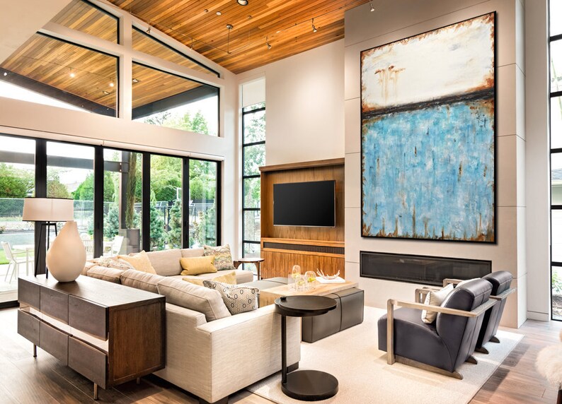 May include: A modern living room with a large abstract painting above a fireplace. The painting features a white background with a blue and brown horizontal stripe. The room has a large sectional sofa, a coffee table, and two armchairs.