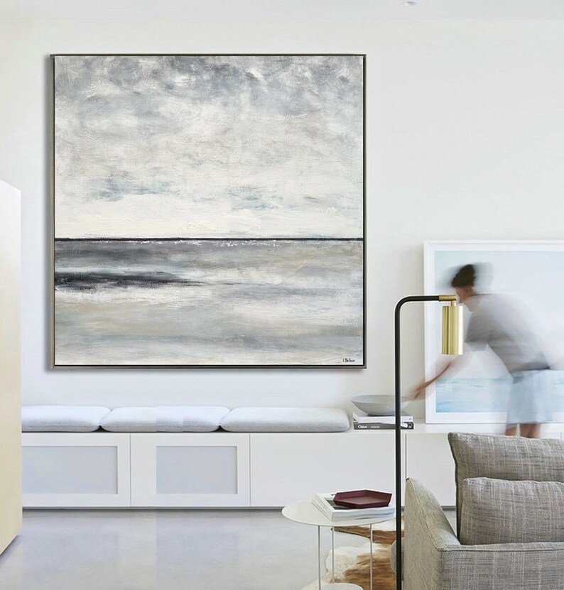 May include: Abstract landscape painting with a gray and white color scheme. The painting depicts a horizon line with a cloudy sky above and a body of water below. The painting is framed in a thin, black frame.