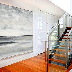 May include: A large abstract painting of a seascape in shades of gray and white. The painting is hanging on a white wall in a modern home with a glass staircase in the background.