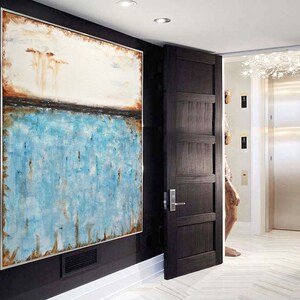 May include: Abstract painting with a white, brown, and blue color scheme. The painting is framed and hanging on a black wall in a hallway.