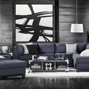 May include: A dark gray sectional sofa with a white and black geometric throw pillow. The sofa is in a living room with a black and white abstract painting on the wall and a coffee table with books and a decorative antler.