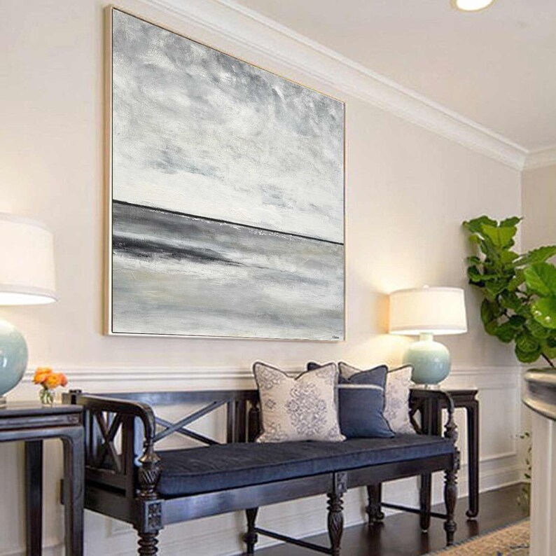 May include: A large abstract painting in shades of gray and white, depicting a seascape with a horizon line and clouds. The painting is hanging on a white wall above a dark wooden bench with blue cushions.
