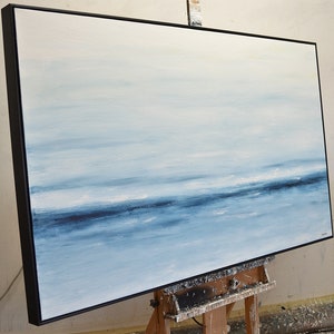 May include: Abstract painting of a seascape with a horizon line and blue and white hues. The painting is framed in a black frame and is on an easel.