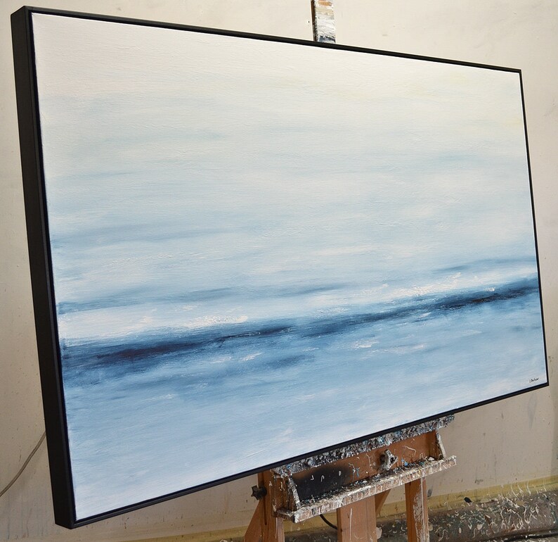 May include: Abstract painting of a seascape with a horizon line and blue and white hues. The painting is framed in a black frame and is on an easel.