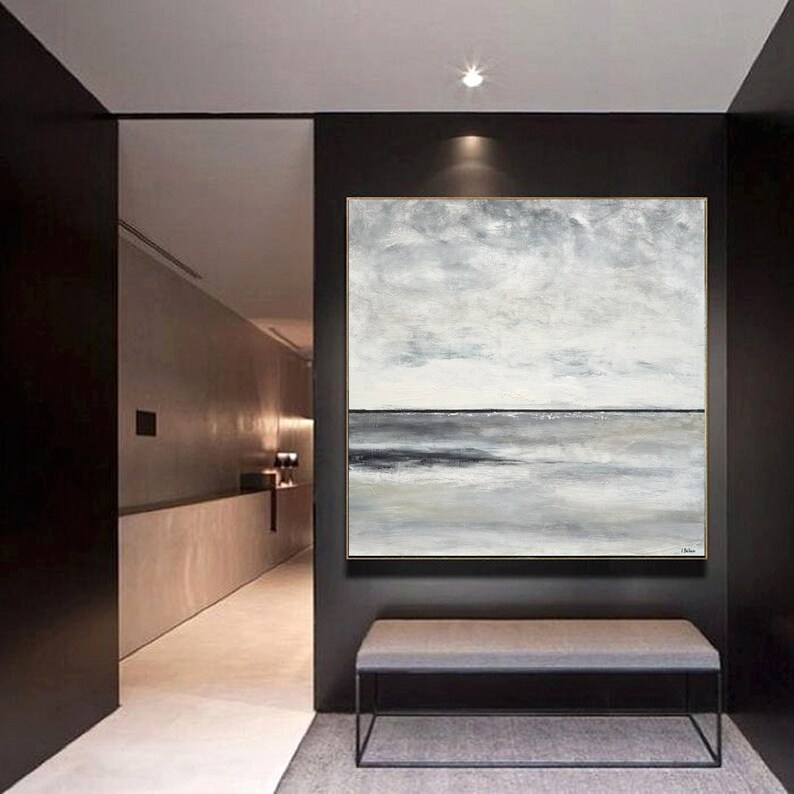 May include: Abstract painting with a gray and white color scheme depicting a seascape. The painting is hanging on a black wall in a hallway with a gray bench underneath.