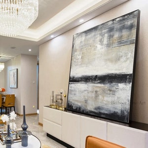 May include: Large abstract painting with a black frame. The artwork features a textured surface with a blend of white, gray, and black hues. The painting is displayed in a modern living space, leaning against a white cabinet.