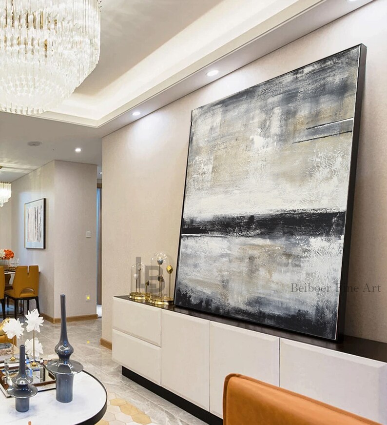 May include: Large abstract painting with a black frame. The artwork features a textured surface with a blend of white, gray, and black hues. The painting is displayed in a modern living space, leaning against a white cabinet.