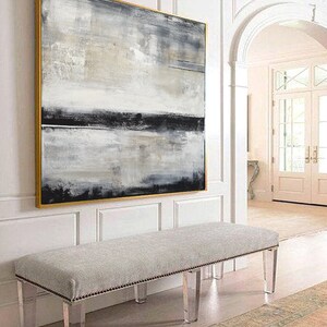 May include: Abstract art painting in a gold frame, featuring a blend of gray, black, and white brushstrokes. A gray upholstered bench with clear acrylic legs sits below the artwork. The scene is set in a well-lit hallway.
