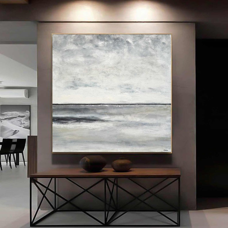May include: Abstract painting of a seascape with a horizon line and gray and white clouds. The painting is framed in a gold frame and is hanging on a wall above a wooden table.