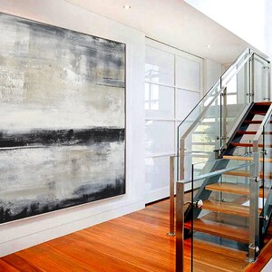 May include: Large abstract painting with a white frame. The artwork features a blend of gray, black, and beige tones, creating a textured, layered effect. The painting hangs on a white wall in a modern interior with a wooden floor and a glass staircase.