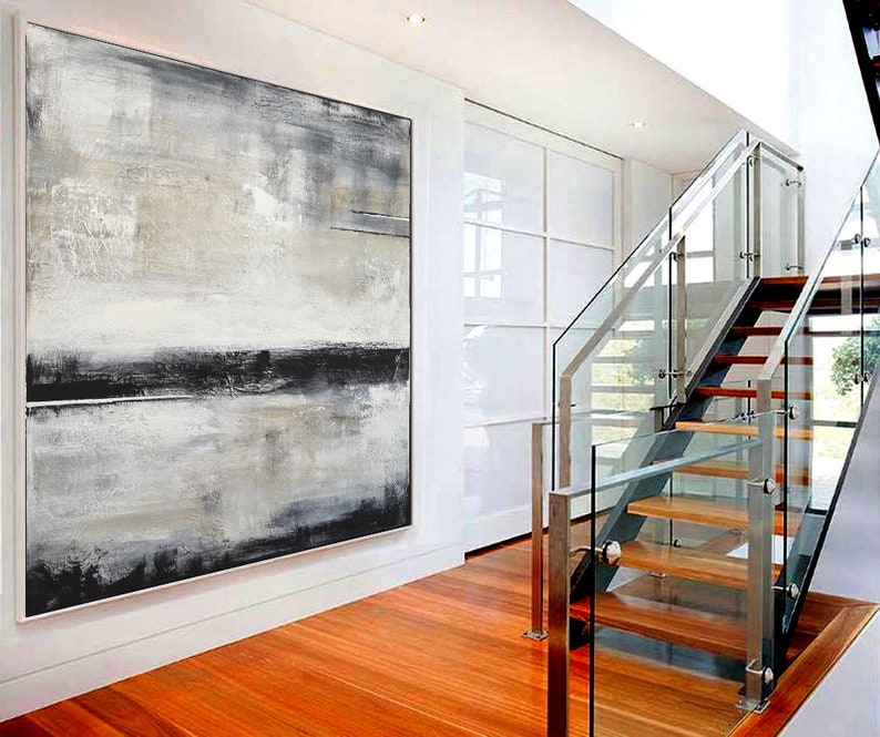 May include: Large abstract painting with a white frame. The artwork features a blend of gray, black, and beige tones, creating a textured, layered effect. The painting hangs on a white wall in a modern interior with a wooden floor and a glass staircase.