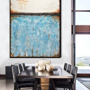 May include: Abstract painting with a white background, a thin black line, and a blue and brown textured bottom. The painting is framed in black and hanging on a white wall above a dining table with chairs.