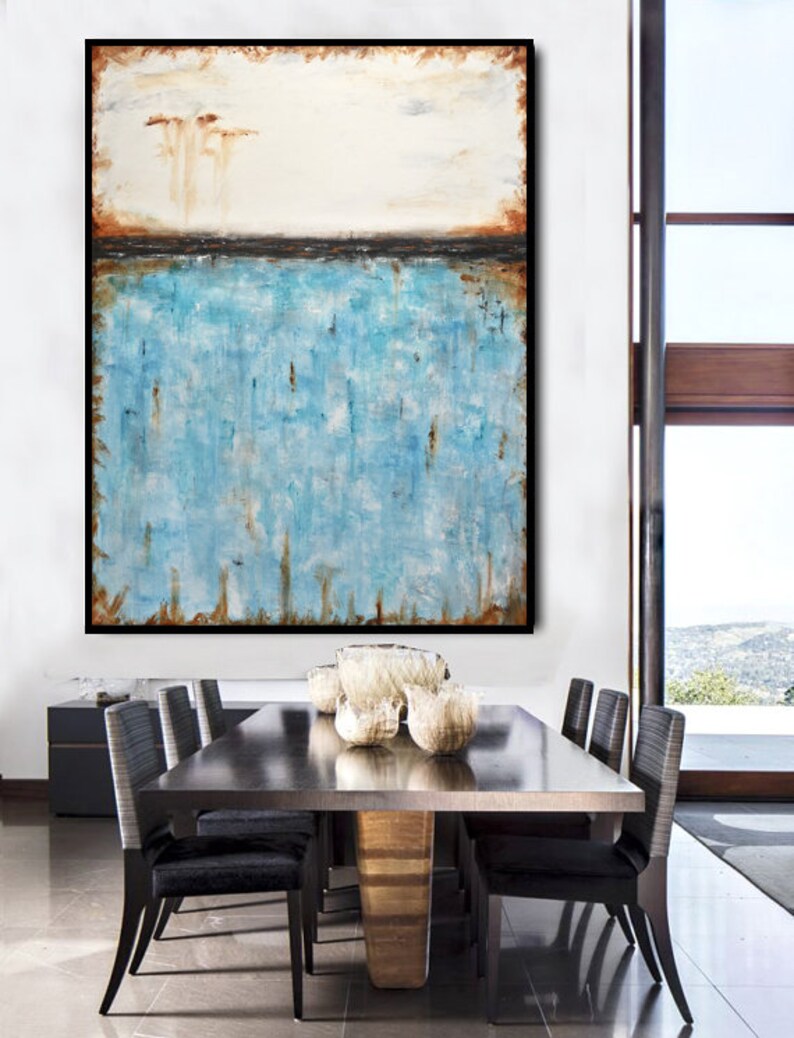 May include: Abstract painting with a white background, a thin black line, and a blue and brown textured bottom. The painting is framed in black and hanging on a white wall above a dining table with chairs.