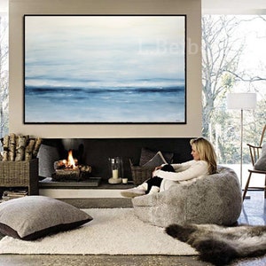 May include: A woman relaxes in a large, fluffy gray bean bag chair in front of a fireplace. A large abstract painting of a blue and white seascape hangs above the fireplace. The room has a cozy and comfortable atmosphere.
