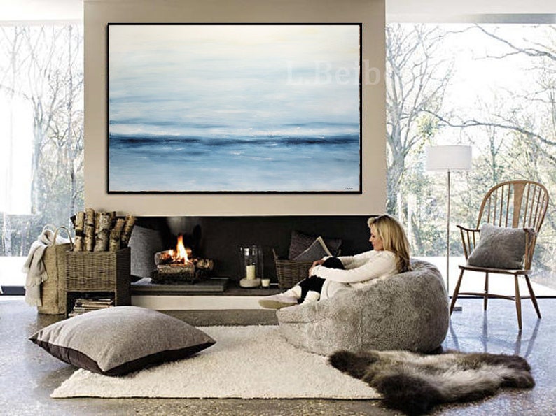 May include: A woman relaxes in a large, fluffy gray bean bag chair in front of a fireplace. A large abstract painting of a blue and white seascape hangs above the fireplace. The room has a cozy and comfortable atmosphere.