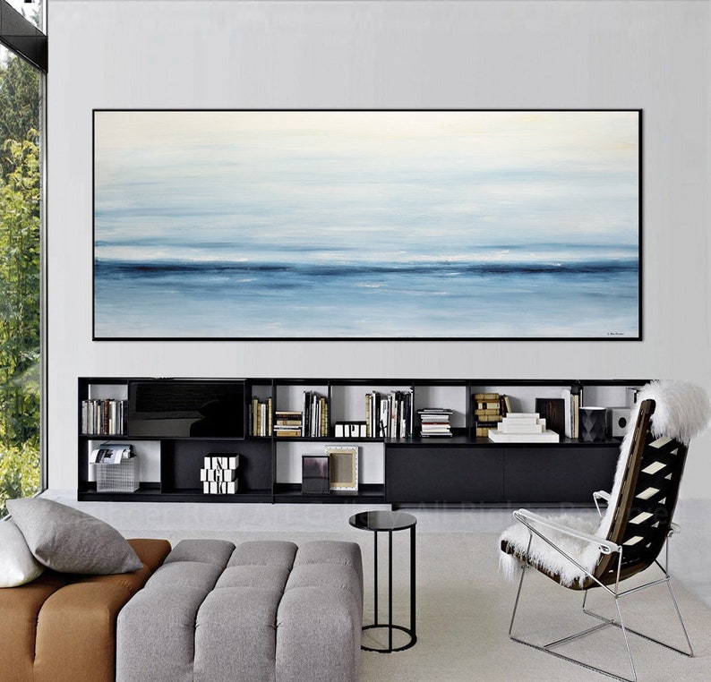 May include: A large abstract painting of a seascape in shades of blue and white hangs above a black bookshelf in a modern living room. The painting is framed in black and has a subtle texture.