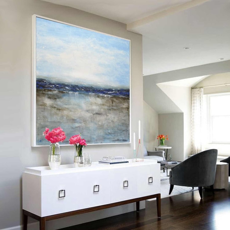May include: A large abstract painting of a seascape with blue and gray tones. The painting is hanging above a white dresser with four drawers. The dresser has a brown wood base and is sitting on a dark wood floor.