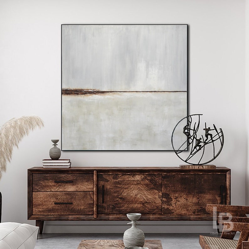 May include: A minimalist abstract painting in shades of gray and brown with a thin horizontal line across the center. The painting is framed in black and hangs above a dark wood dresser with two drawers and a cabinet door. A black metal sculpture of figures in motion sits on the dresser.