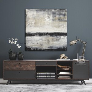 May include: Abstract square painting in neutral tones of gray, black, and beige hangs above a dark wood and gray cabinet. The cabinet has drawers and open shelves. A metal desk lamp and decorative items sit on the cabinet.