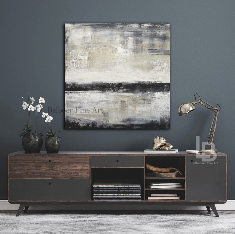 May include: Abstract square painting in neutral tones of gray, black, and beige hangs above a dark wood and gray cabinet. The cabinet has drawers and open shelves. A metal desk lamp and decorative items sit on the cabinet.