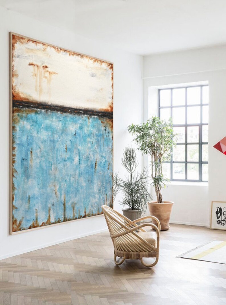 May include: Abstract painting with a blue, white, and brown color scheme. The painting is hanging on a white wall in a room with a wooden floor and a wicker chair.