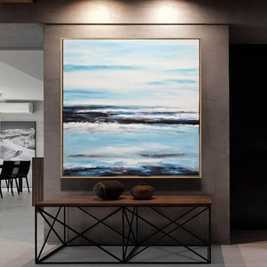 Pu&ograve; includere: Dipinto astratto di un paesaggio marino con tonalit&agrave; blu, bianche e nere. Il dipinto &egrave; incorniciato da una cornice dorata e appeso a una parete grigia sopra un tavolo di legno con una base in metallo nero.