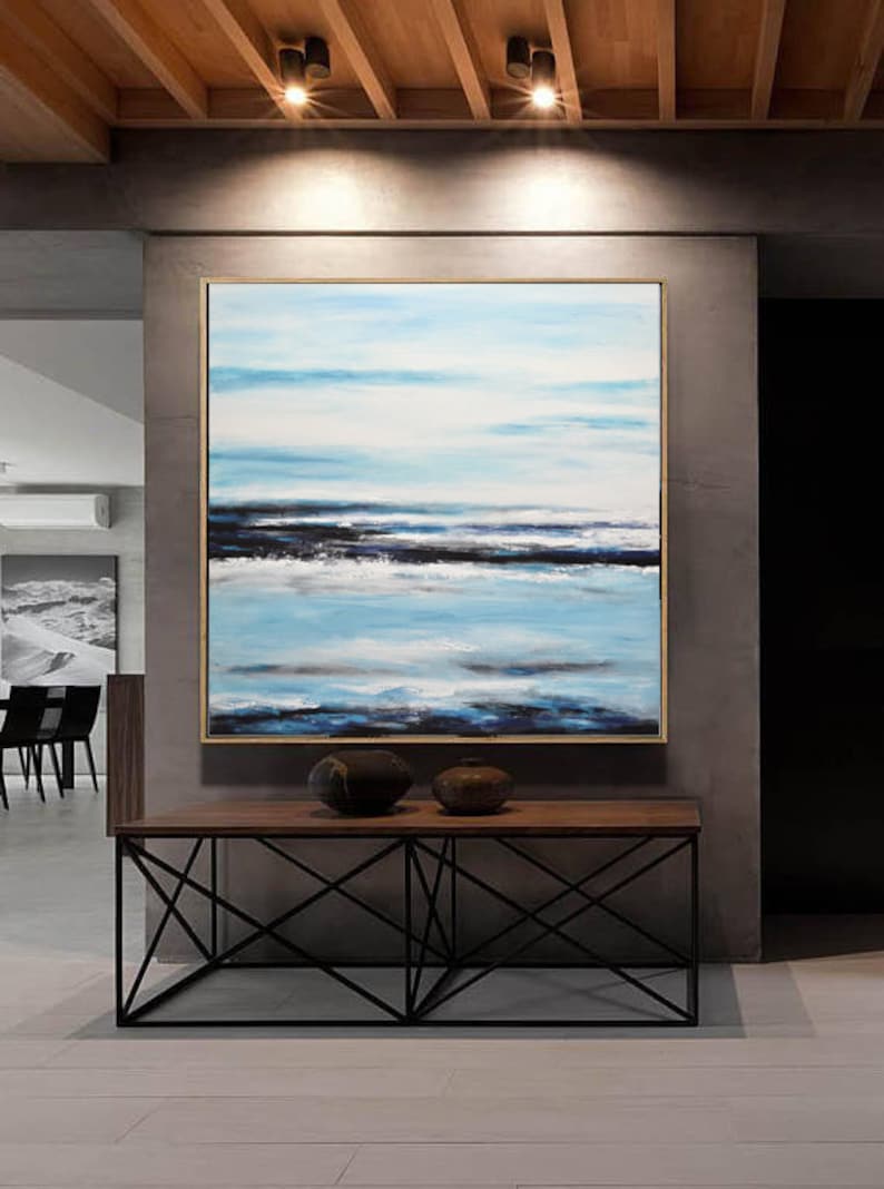 Pu&ograve; includere: Dipinto astratto di un paesaggio marino con tonalit&agrave; blu, bianche e nere. Il dipinto &egrave; incorniciato da una cornice dorata e appeso a una parete grigia sopra un tavolo di legno con una base in metallo nero.