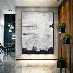 Op de afbeelding: Groot abstract schilderij in een modern interieur. Het kunstwerk heeft een textuur, monochroom ontwerp met tinten wit, grijs en zwart. Het schilderij is ingelijst en aan een lichtgrijze muur bevestigd. De kamer heeft een minimalistisch ontwerp met planten.