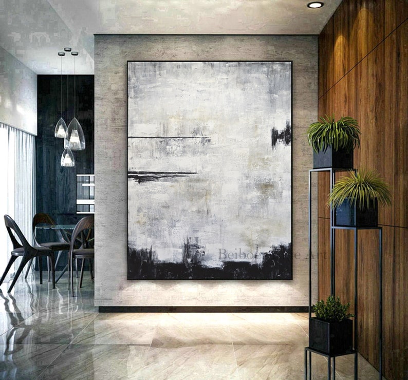 Op de afbeelding: Groot abstract schilderij in een modern interieur. Het kunstwerk heeft een textuur, monochroom ontwerp met tinten wit, grijs en zwart. Het schilderij is ingelijst en aan een lichtgrijze muur bevestigd. De kamer heeft een minimalistisch ontwerp met planten.