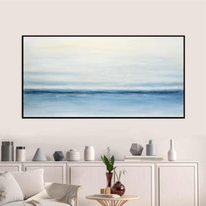 May include: Abstract painting of a seascape with a blue horizon and white clouds. The painting is framed in black and hangs on a white wall above a white shelf with decorative objects.