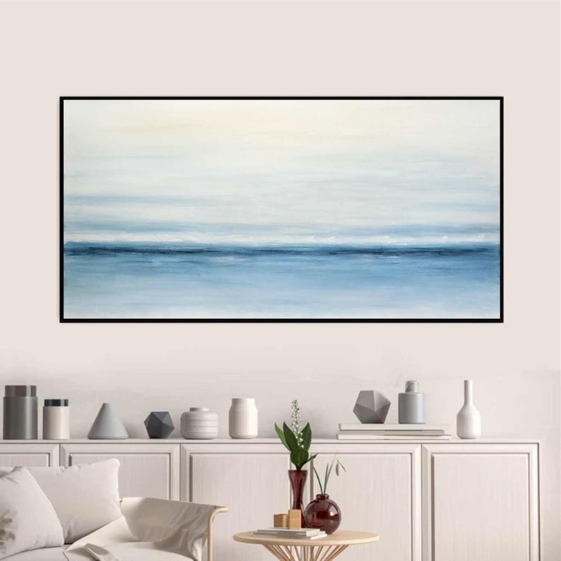 May include: Abstract painting of a seascape with a blue horizon and white clouds. The painting is framed in black and hangs on a white wall above a white shelf with decorative objects.