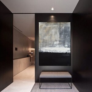 May include: A large abstract painting in shades of gray, white, and black hangs on a black wall in a modern hallway. The painting features a horizontal band of white across the middle, with a darker gray area above and a black area below. The painting is framed by a black wall and a white ceiling.