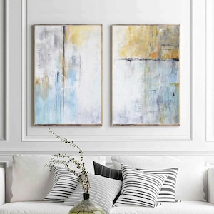 May include: Two abstract paintings in a set. The paintings are in shades of blue, yellow, and gray. The paintings are framed in a simple, light wood frame.