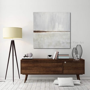 May include: A square abstract painting with a horizontal brown line across the middle. The painting is on a white wall above a dark brown wooden dresser with two drawers. The dresser is in a room with a white floor and a white lamp with a cream-colored shade.