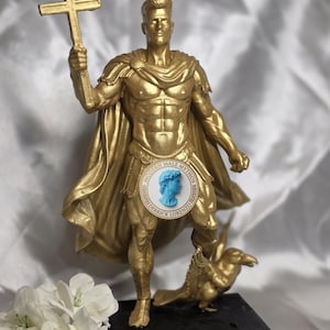 Peut inclure: Une statue dorée représentant une silhouette musclée portant une cape et tenant une croix. Un petit oiseau se tient aux pieds de la silhouette sur un socle noir. Un emblème circulaire avec un profil bleu et le texte MODERN MALE MYTHOS DERRICK LORENZO est centré.