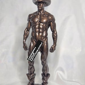 May include: A bronze-colored statue of a muscular figure wearing a cowboy hat and boots. The statue has a Modern Male Mythos.com banner covering the lower torso. The figure is standing on a small, dark base.