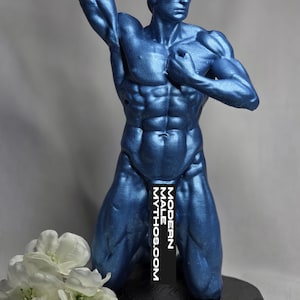 May include: A blue sculpture of a muscular male figure in a kneeling pose, with one arm raised above the head. The sculpture is on a black base, and the text "MODERN MALE MYTHOS.COM" is visible. White flowers are in the foreground.