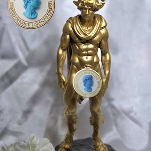 Puede incluir: Una estatua de color dorado de una figura de sátiro con cuernos y patas de cabra, sobre una base oscura. La figura lleva una tela drapeada alrededor del cuello. Es visible un logotipo circular con un busto azul y el texto MODERN MALE MYTHOS DERRICK LORENZO.