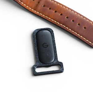May include: A black watch strap holder with the Google logo, next to a brown leather watch band with white stitching. The watch band has several holes for adjustment. The background is white.