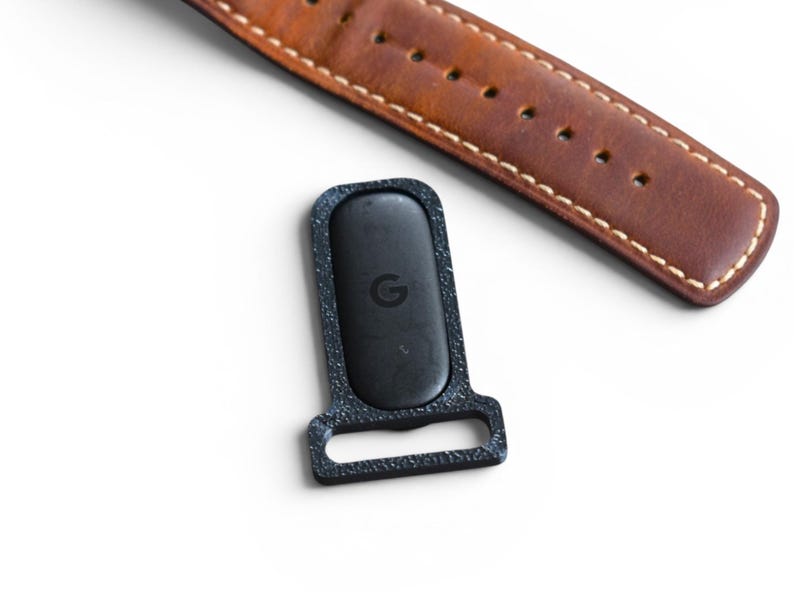May include: A black watch strap holder with the Google logo, next to a brown leather watch band with white stitching. The watch band has several holes for adjustment. The background is white.