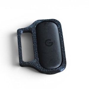 May include: A black, rectangular device with a textured, speckled frame and a smooth, oval-shaped center. The center features a small "G" logo. The device has a handle-like extension on one side.