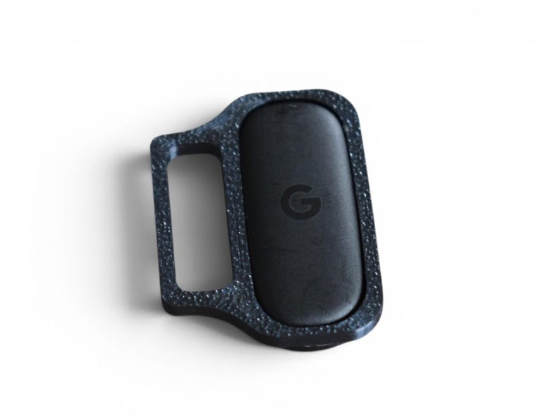 May include: A black, rectangular device with a textured, speckled frame and a smooth, oval-shaped center. The center features a small "G" logo. The device has a handle-like extension on one side.