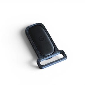 May include: A black rectangular object with a rounded top and a loop at the bottom. The object has a textured, dark gray border and a black center with a small, circular emblem. The item is set against a white background.