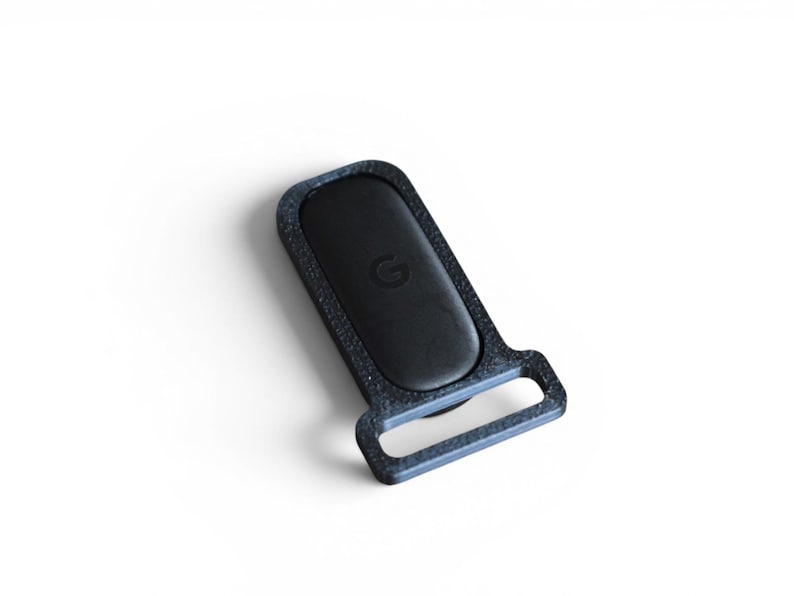 May include: A black rectangular object with a rounded top and a loop at the bottom. The object has a textured, dark gray border and a black center with a small, circular emblem. The item is set against a white background.